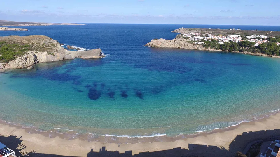 Enseada ampla em arco de Arenal d'en Castell, com praia e edifícios na costa.