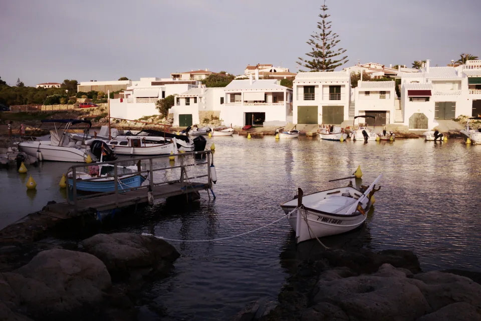 Casas brancas de Binibeca Vell junto à água, com embarcações e rochas em primeiro plano.