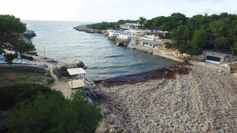 Costa rochosa e arenosa de Cala Blanca, com árvores e estruturas junto à água.