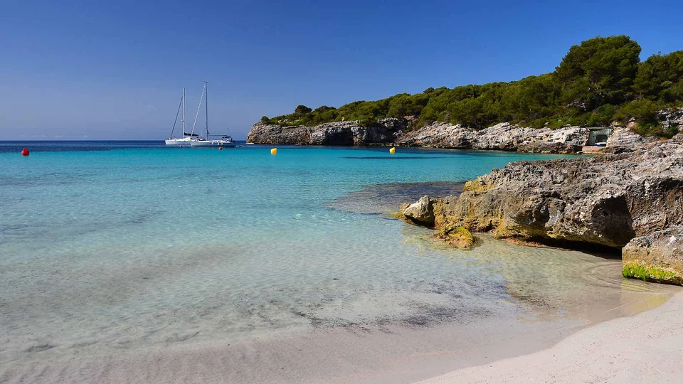 Margem de areia e água clara em Cala en Turqueta, entre rochas e pinheiros.
