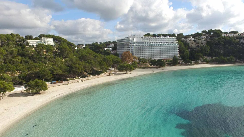 A baía ampla de Cala Galdana, com praia, edifícios visíveis e pinheiros na encosta.