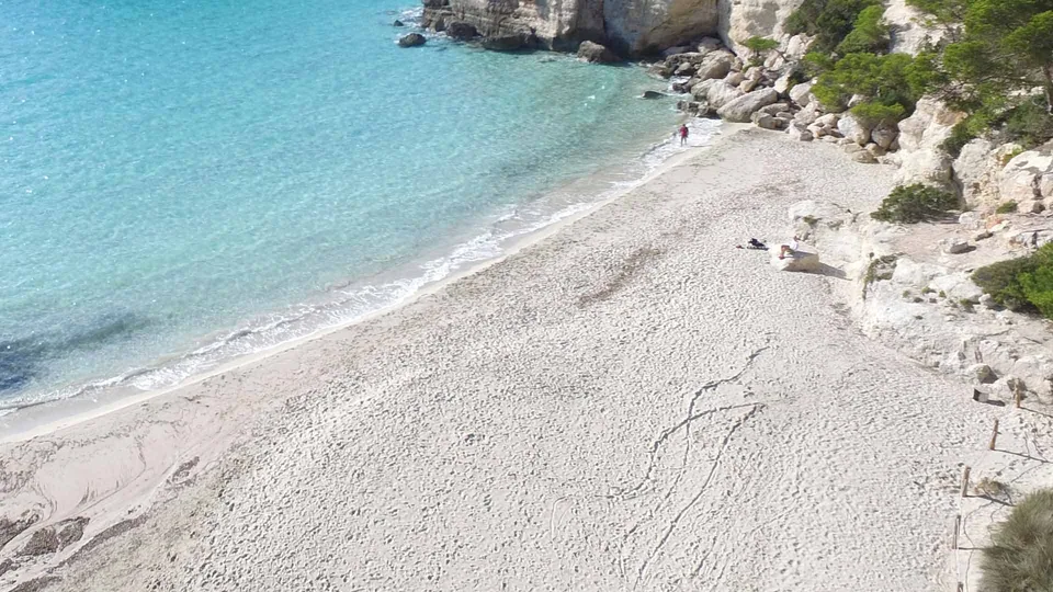 A praia clara de Cala Mitjana junto à água límpida e à orla rochosa.