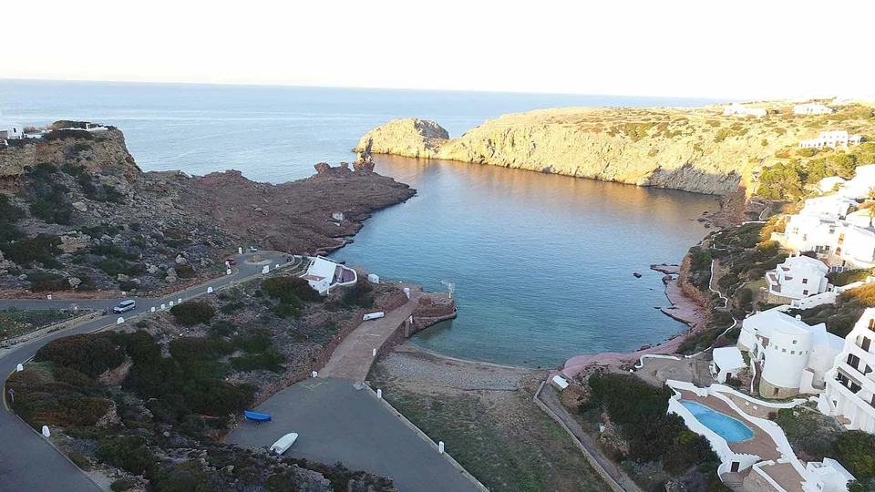 Enseada de Cala Morell entre promontórios rochosos, com edifícios brancos nas encostas.