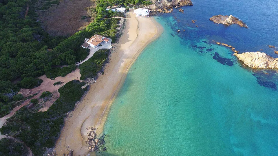 Enseada arenosa de Cala Pregonda, com água turquesa, rochas e margem arborizada.