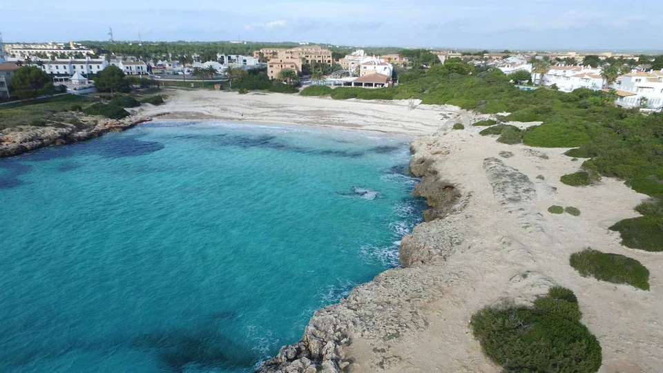 Praia de Cala en Bosch, com areia entre margens rochosas e edifícios da urbanização ao fundo.