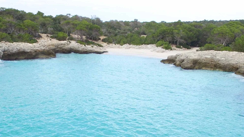 Pequena enseada turquesa de Es Talaier, entre rocha clara e vegetação baixa.