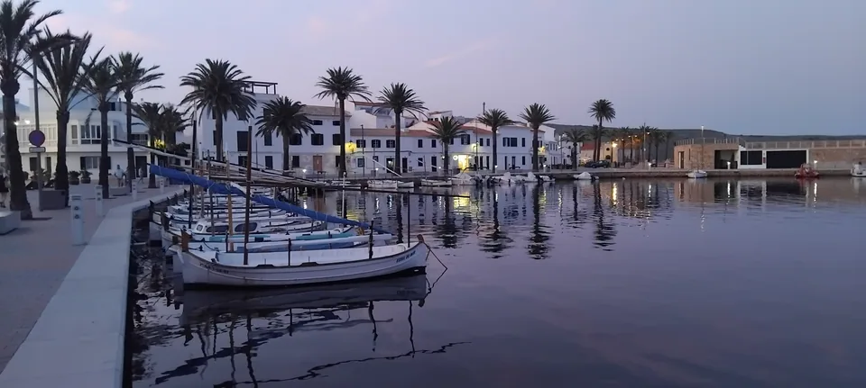 Barcos no porto de Fornells ao entardecer, com palmeiras e edifícios junto ao cais.