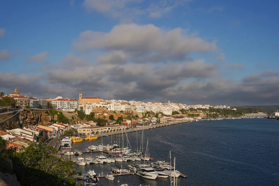 Vista ampla do porto de Maó, com barcos e frentes edificadas junto à água.