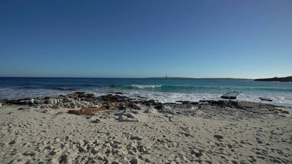 Costa rochosa e mar aberto na praia de Punta Prima, com uma forma pequena ao longe.